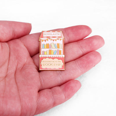 The "Scholastic Book Fair" Enamel Pin features a stylised bookshelf in lovely pastel shades of pink, coral, and gold, commemorating everyone's favourite childhood memory: The Scholastic Book Fair! Oh, the joy! So many books!