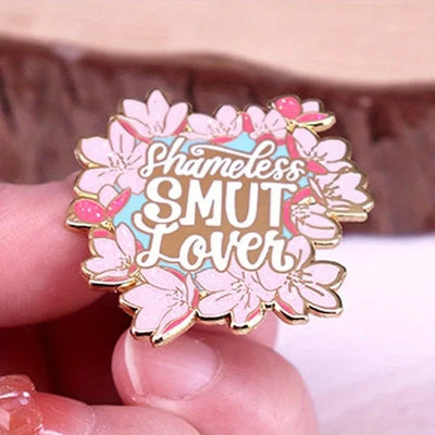 The "Shameless Smut Lover" Enamel Pin features a saucy declaration of your love for naughty romance novels, surrounded by a ring of cute cherry blossoms.