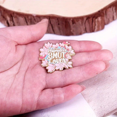 The "Shameless Smut Lover" Enamel Pin features a saucy declaration of your love for naughty romance novels, surrounded by a ring of cute cherry blossoms.