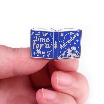 The "Time For A New Adventure" Enamel Pin features a small blue and silver book lying open, with the words "time for a new adventure" scrawled across the front and back cover, along with small illustrations of mountains and trees.