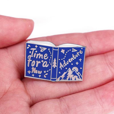 The "Time For A New Adventure" Enamel Pin features a small blue and silver book lying open, with the words "time for a new adventure" scrawled across the front and back cover, along with small illustrations of mountains and trees.