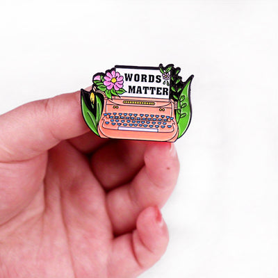 The "Words Matter" Enamel Pin features a stylised typewriter adorned with flowers and leaves, with "Words Matter" printed on the typewriter's paper. So true! They really do!