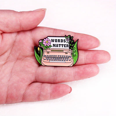 The "Words Matter" Enamel Pin features a stylised typewriter adorned with flowers and leaves, with "Words Matter" printed on the typewriter's paper. So true! They really do!