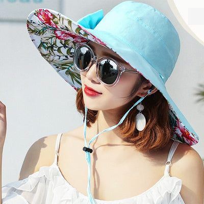 Caribbean Cruise Reversible Sunhat - A large-brimmed, floppy sunhat with a solid colour on one side and a vibrant floral print on the other, which can be worn either way depending on the user's choice. It is decorated with a large removable bow. This image shows a model wearing the Blue colour, which is a very soft baby blue, almost a cyan.