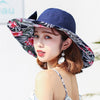 Caribbean Cruise Reversible Sunhat - A large-brimmed, floppy sunhat with a solid colour on one side and a vibrant floral print on the other, which can be worn either way depending on the user's choice. It is decorated with a large removable bow. This image shows a model wearing the Navy colour, which is a very dark blue. The floral print also has a dark blue background.