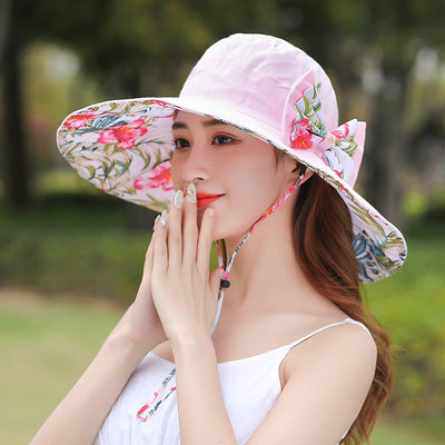Caribbean Cruise Reversible Sunhat - A large-brimmed, floppy sunhat with a solid colour on one side and a vibrant floral print on the other, which can be worn either way depending on the user's choice. It is decorated with a large removable bow. This image shows a model wearing the Pink colour, which is a very soft pastel pink shade.