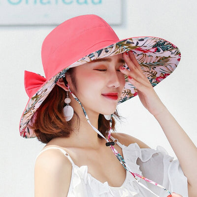 Caribbean Cruise Reversible Sunhat - A large-brimmed, floppy sunhat with a solid colour on one side and a vibrant floral print on the other, which can be worn either way depending on the user's choice. It is decorated with a large removable bow. This image shows a model wearing the Salmon colour, which is a vibrant and highly-saturated shade of orangey-pink.